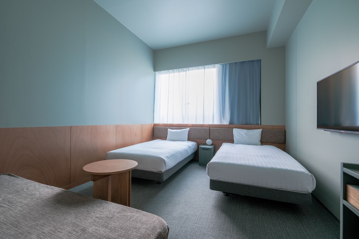 A hotel room with two beds, a small round table, a TV mounted on the wall, and a window with curtains.