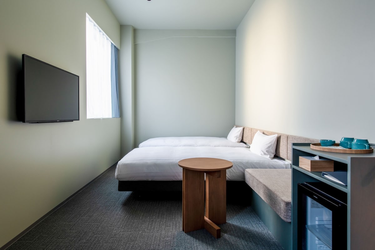 A small, modern hotel room with a bed, a TV, a small table, and a minibar.