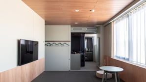 A modern hotel room with a flat-screen TV, a small kitchenette, a seating area with a round table, and a large window with curtains.
