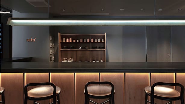 A bar area with a wooden counter, three bar stools, and a shelf with various items.