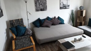 A modern bedroom with a bed, a chair with patterned cushions, a bedside table with a remote control, and two wall-mounted pictures of cities. - Apartment Come-Home (Hohnstedt)
