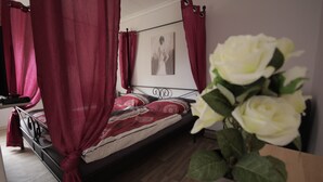 A bedroom with a four-poster bed, red curtains, a framed picture on the wall, and a potted white rose. - Apartment Come-Home (Hohnstedt)