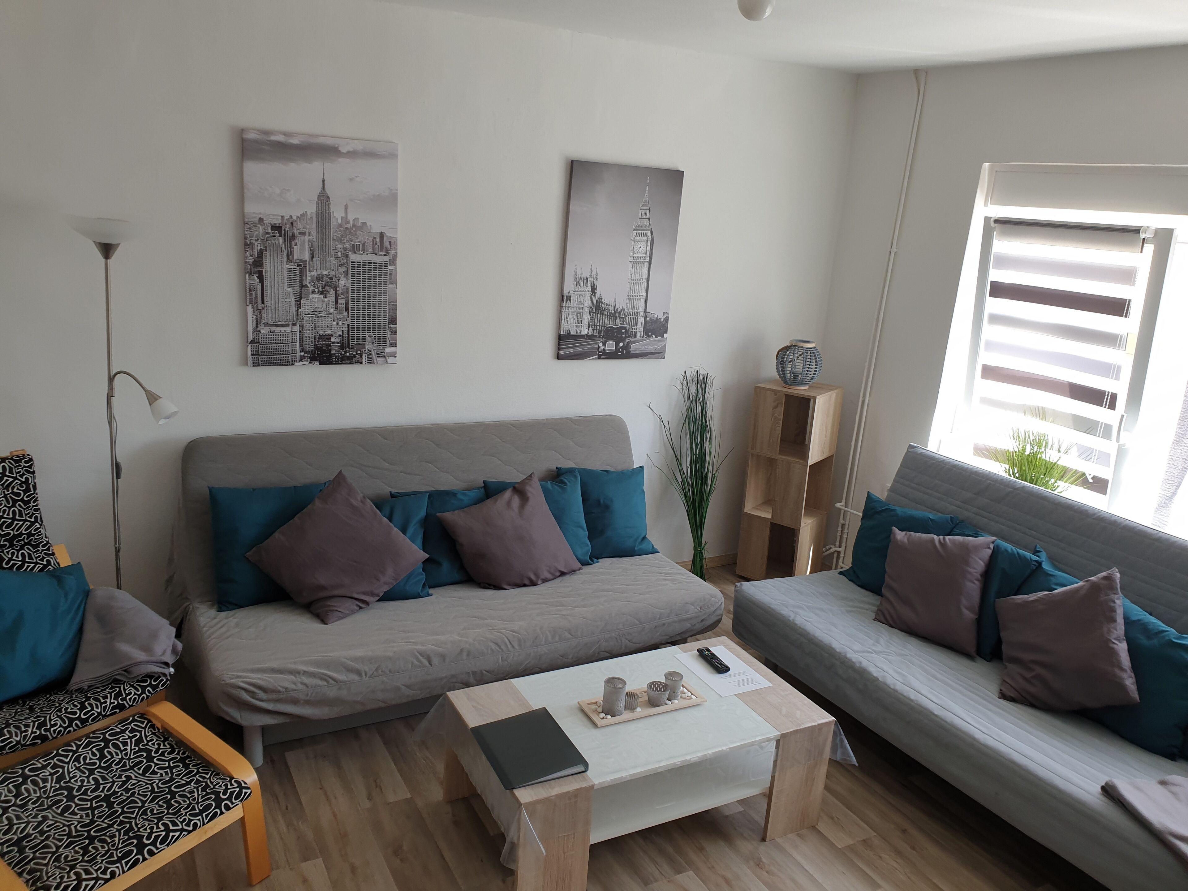 A modern living room with a grey sofa, teal and brown cushions, a wooden coffee table with a remote and decorative items, and two cityscape wall art pieces.