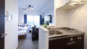 A compact hotel room with a kitchenette, a bed with a headboard, a seating area, and a window with curtains.