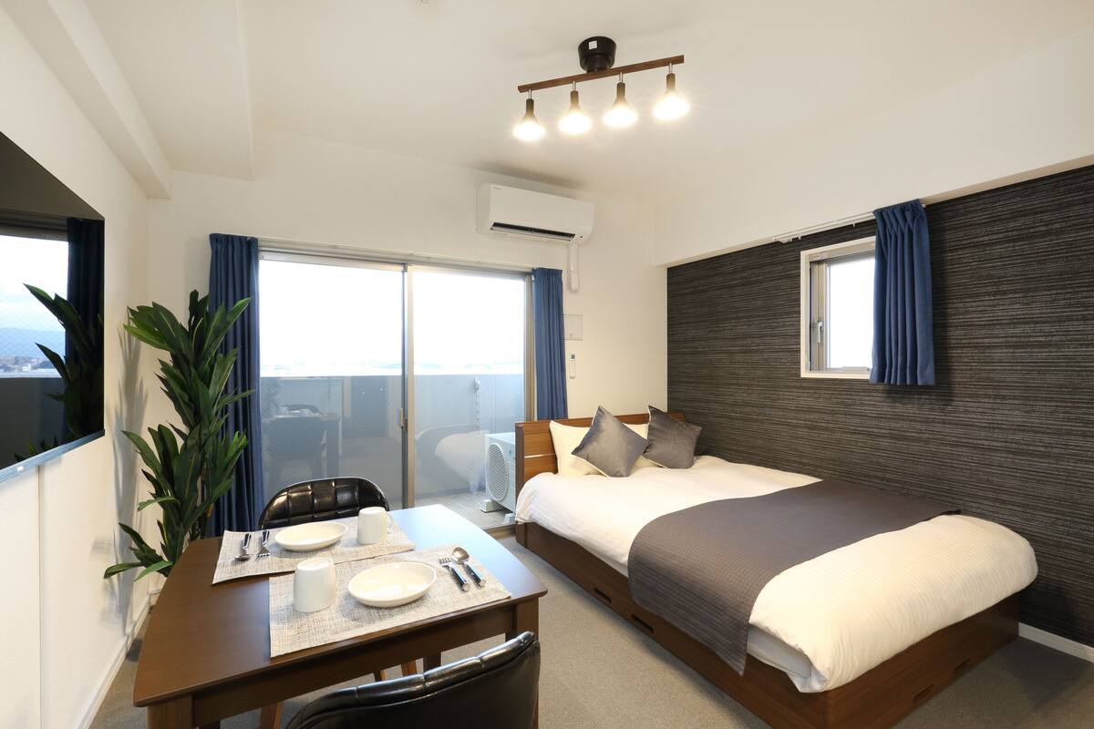 A hotel room with a bed, a dining table with plates and cutlery, a chair, a potted plant, and a window with curtains.