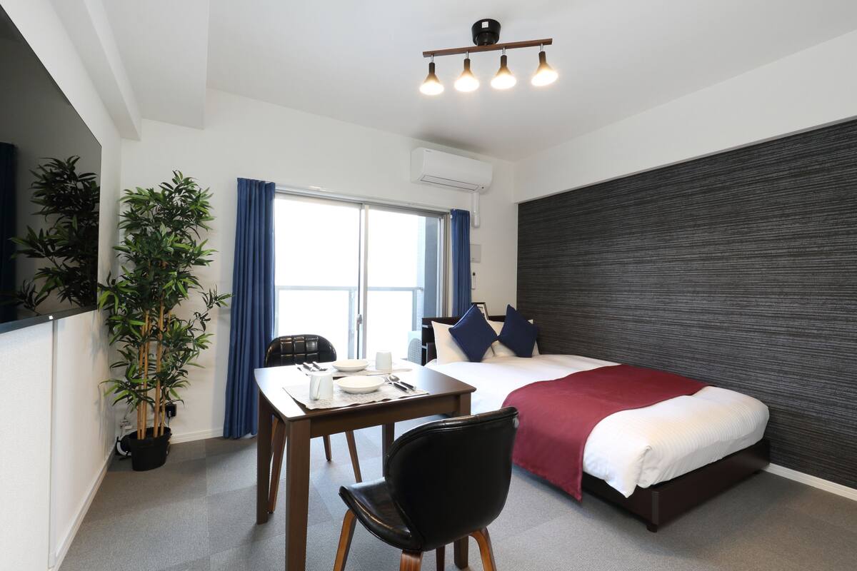 A hotel room with a bed, a desk with a chair, a potted plant, and a window with curtains.