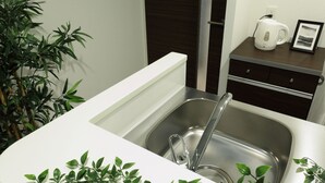 A modern kitchen with a stainless steel sink, a white countertop, and a small cabinet with a kettle and picture frame.