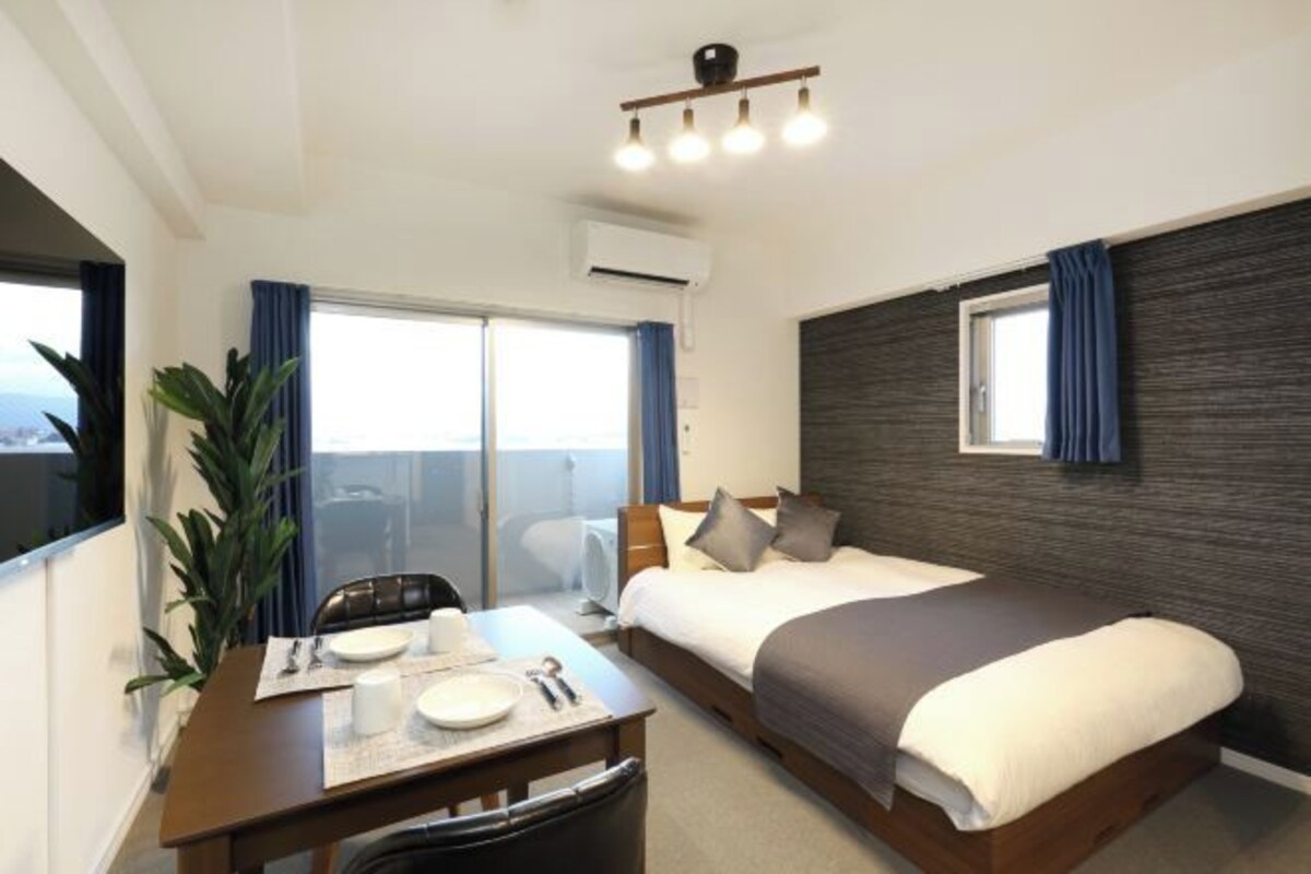 A hotel room with a bed, a desk with a chair, a dining table with plates and cutlery, and a large window with curtains.