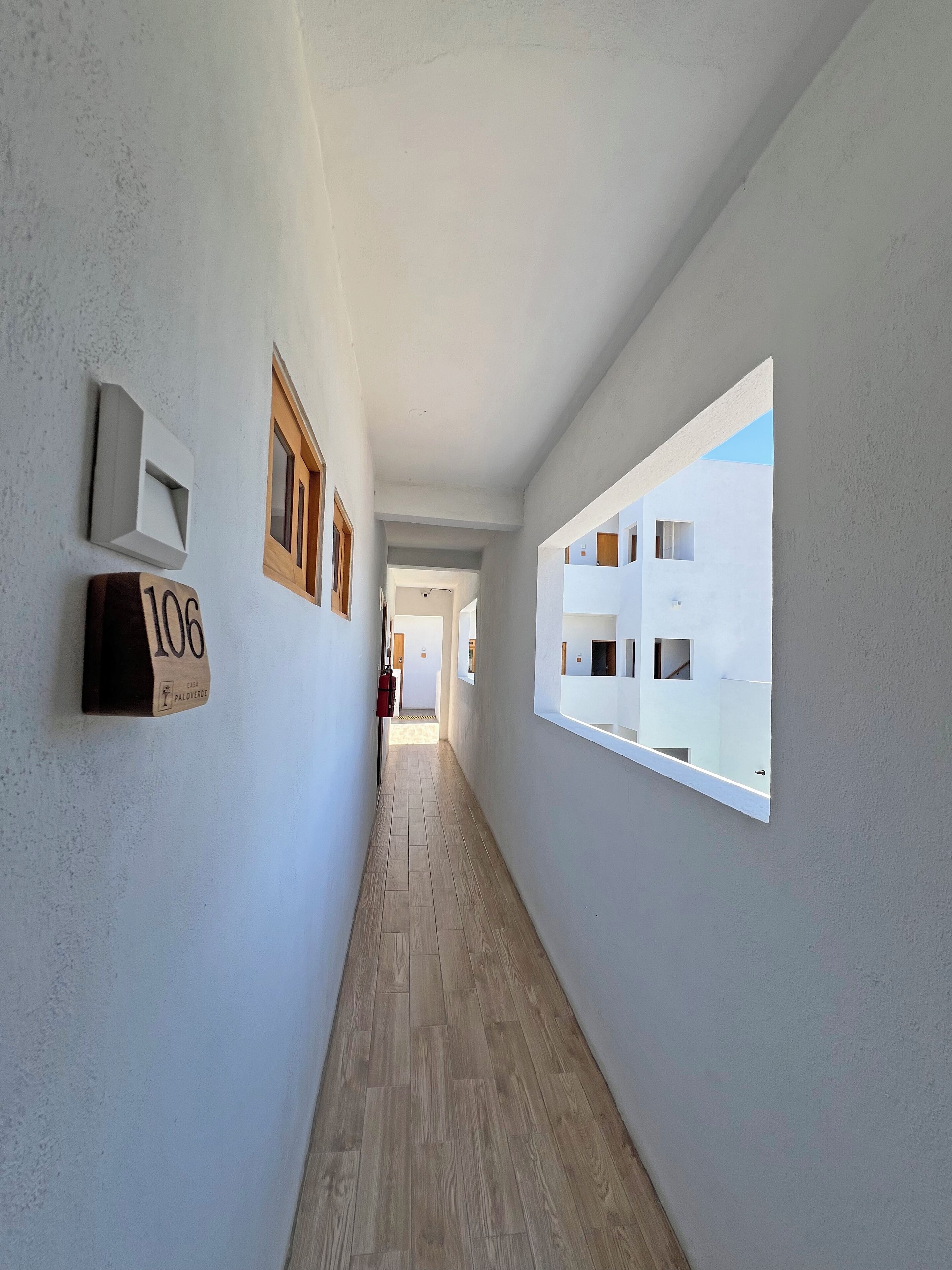 A narrow hallway with wooden flooring, white walls, and a large window on the right. There are two light fixtures and a door at the end.