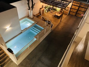 Un área interior de piscina moderna con dos piscinas, una zona de estar con una mesa y sillas, y una escalera que conduce a otro nivel.
