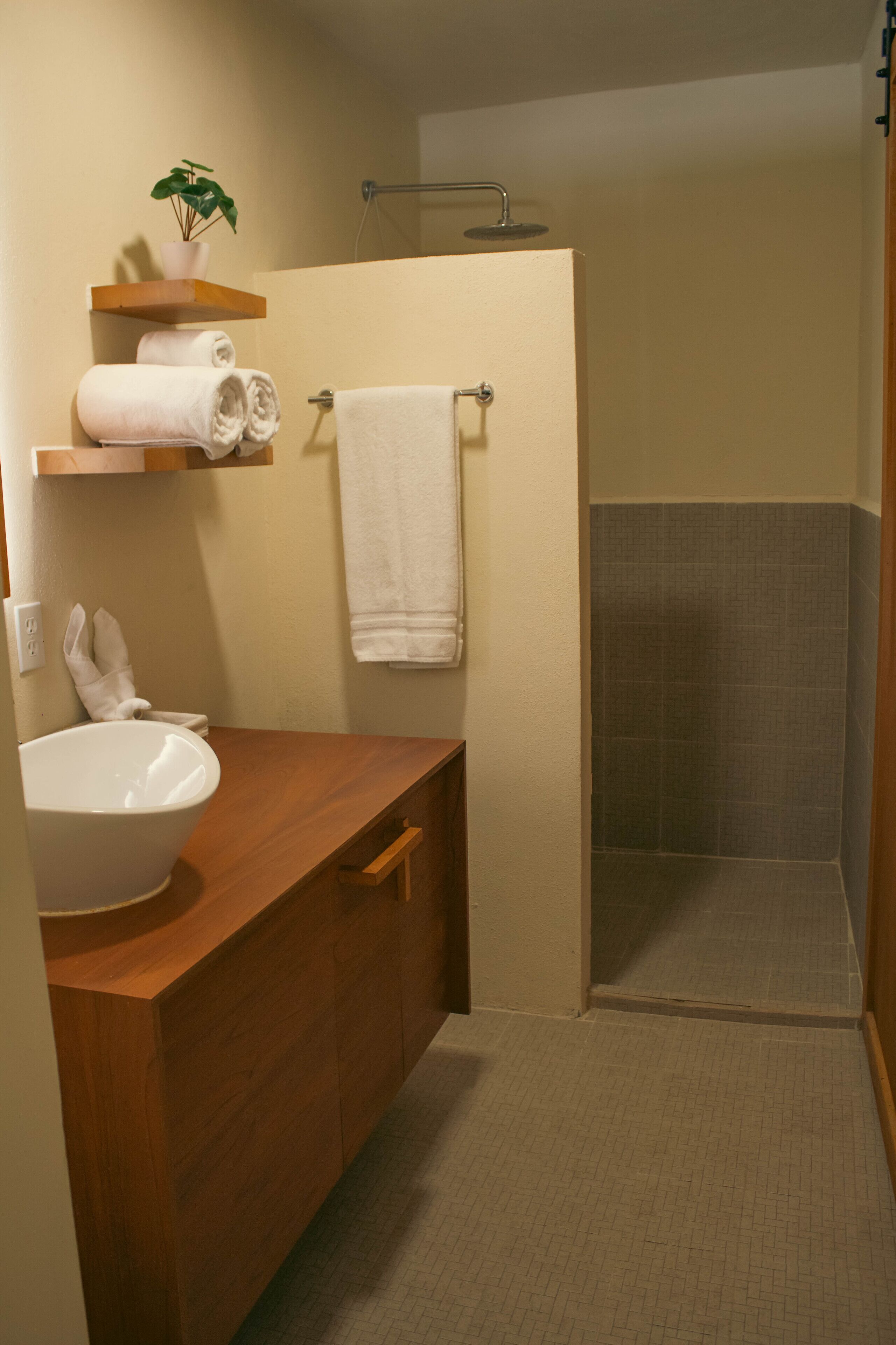 Un baño con tocador de madera, un lavabo blanco, un perchero con toallas, zona de ducha y una pequeña planta sobre una estantería.