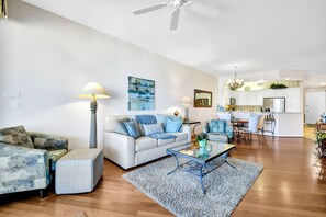 A modern living room with a white sofa, blue accent pillows, a glass coffee table, and a dining area with a chandelier. - Regatta - Enjoy Gulf Views, Walk to the Beach! (Naples)