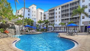 A swimming pool with lounge chairs and umbrellas, surrounded by a building with balconies and a waterfall feature. - Regatta - Enjoy Gulf Views, Walk to the Beach! (Naples)