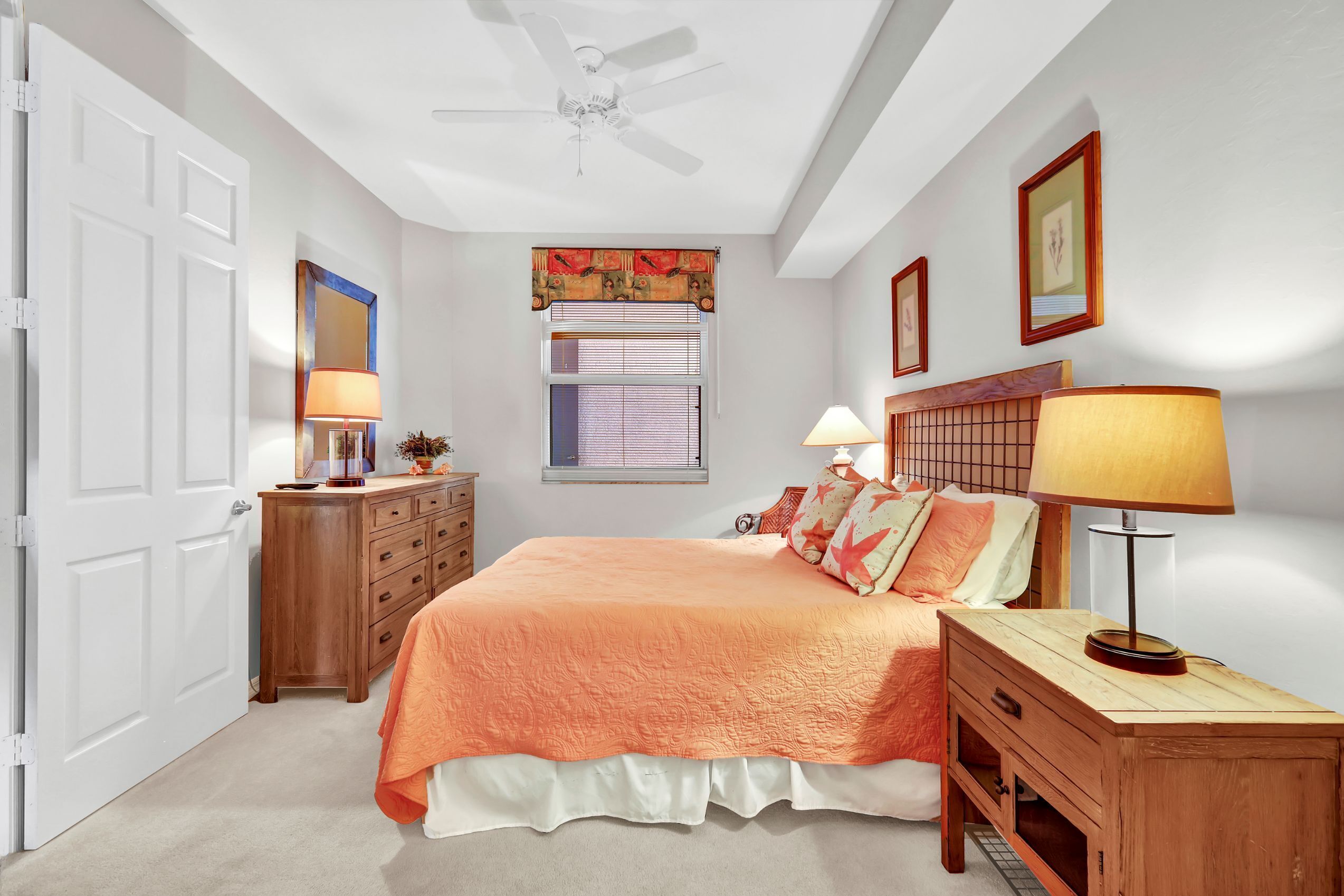 A bedroom with a bed, bedside tables, a dresser, a ceiling fan, a window with blinds, and framed pictures on the wall.