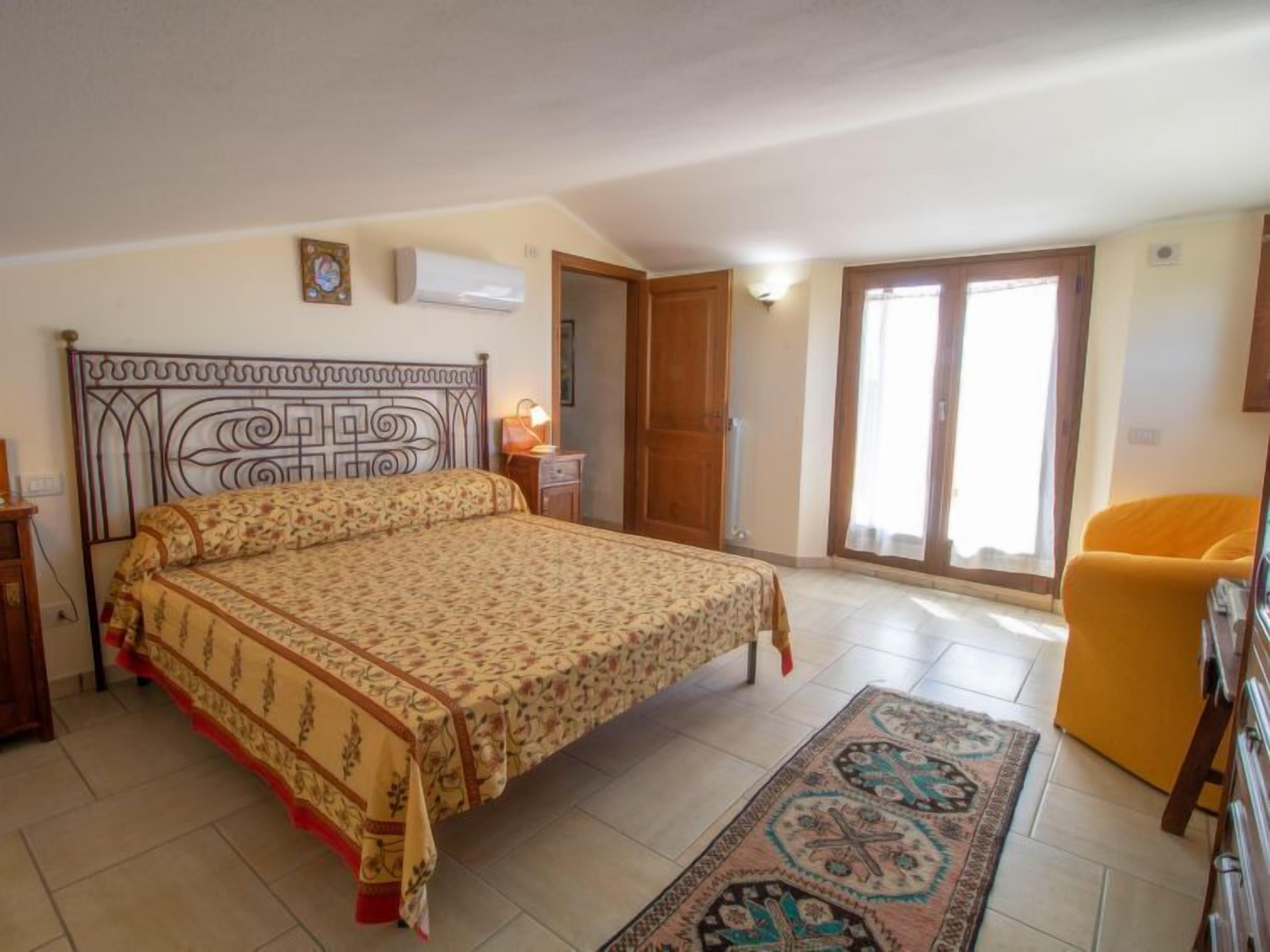 Une chambre à coucher comprenant un lit, une table de chevet, une lampe, une chaise et une porte donnant sur un balcon.