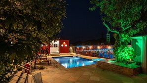 An outdoor swimming pool area with a red building, a tree with a hanging lantern, and a small table with a bottle. - The G Snoozebox (Udaipur)