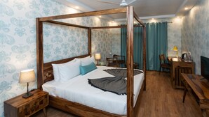A four-poster bed with a wooden frame, a bedside table with a lamp, and a desk with a chair in a room with patterned wallpaper and a ceiling fan. - The G Snoozebox (Udaipur)