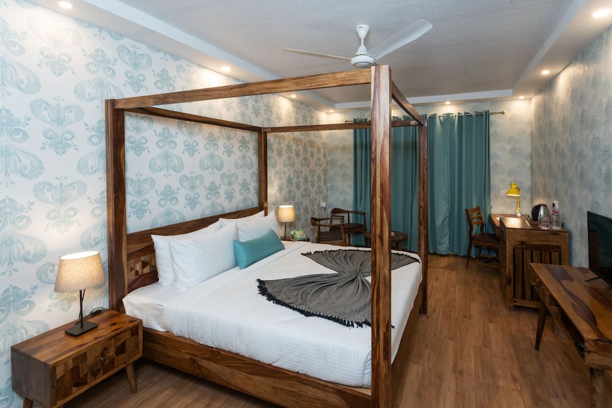 Un letto a baldacchino con struttura in legno, un comodino con lampada, e una scrivania con sedia in una stanza con carta da parati decorata e ventilatore a soffitto.
