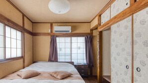 A traditional Japanese-style room with tatami flooring, a bed with a white and brown bedspread, a wooden headboard, and a large window with curtains.