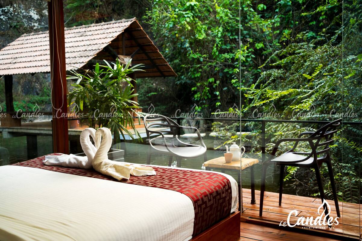 A bedroom with a bed, a glass table, two chairs, and a view of a lush garden.