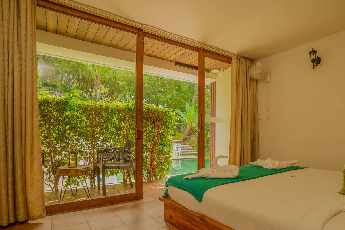 A bedroom with a large bed, a view of a lush garden, and a sliding glass door leading to an outdoor area.