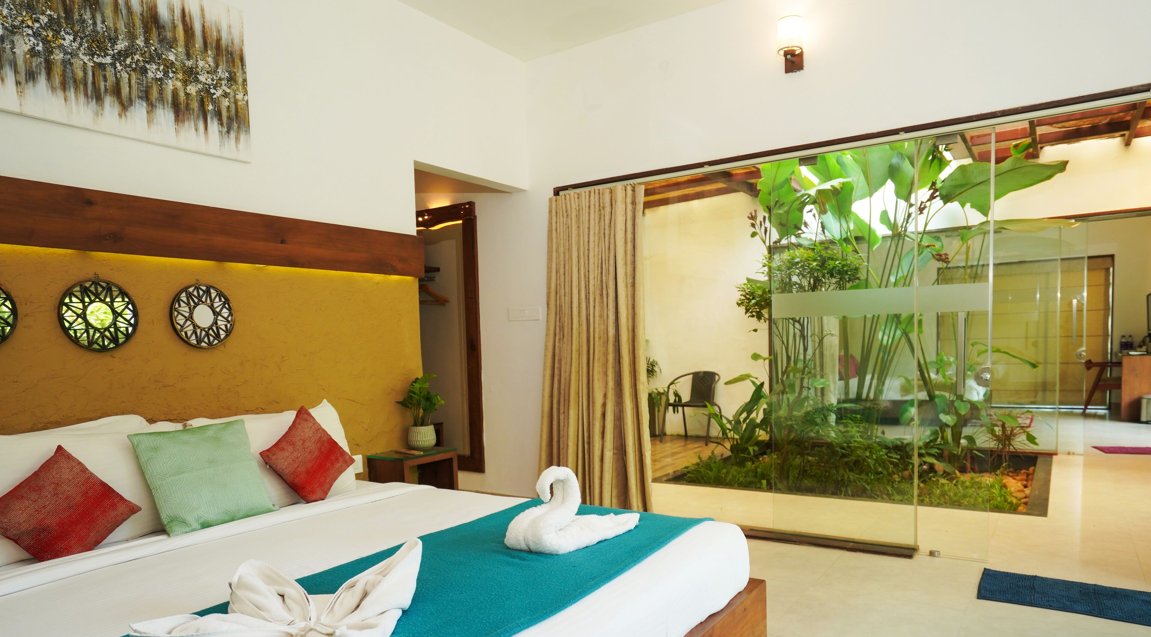 Habitación de hotel con cama, toalla con forma de cisne, puerta de vidrio que da a un balcón con plantas y decoración en la pared.