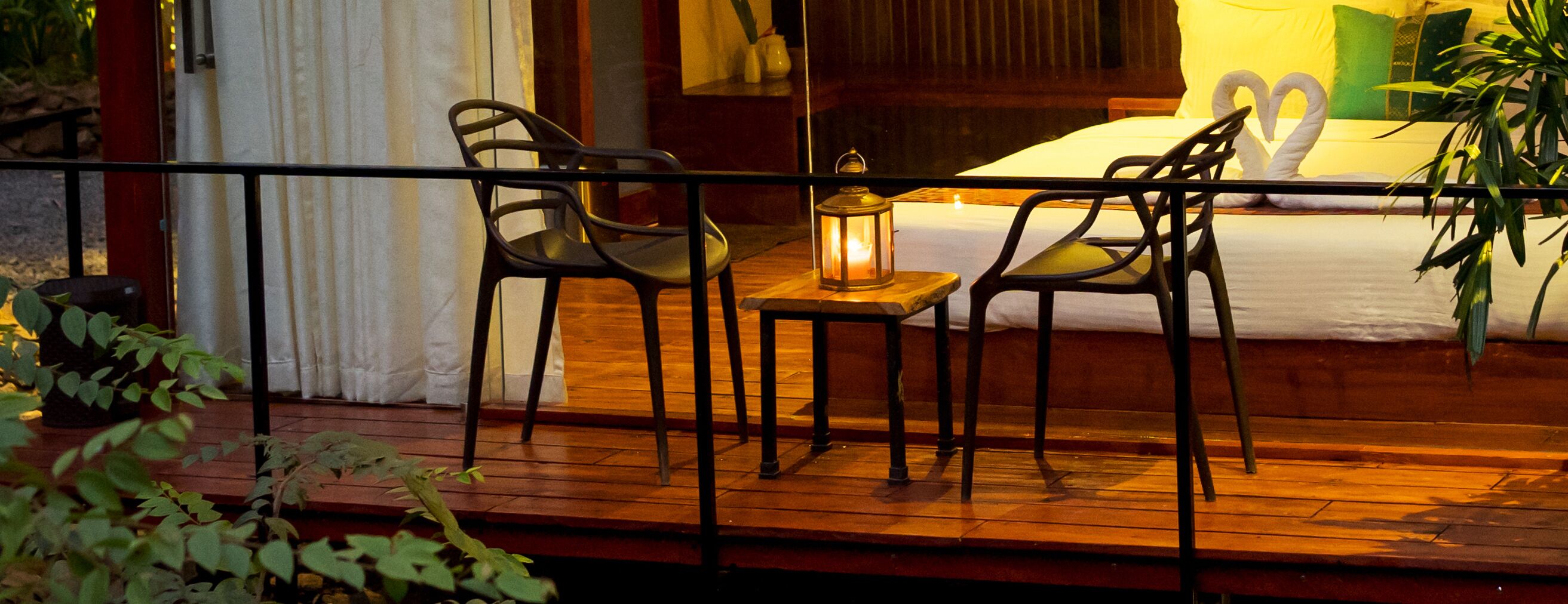 A wooden deck with two chairs and a table, a lantern, and a bed with a swan-shaped pillow.