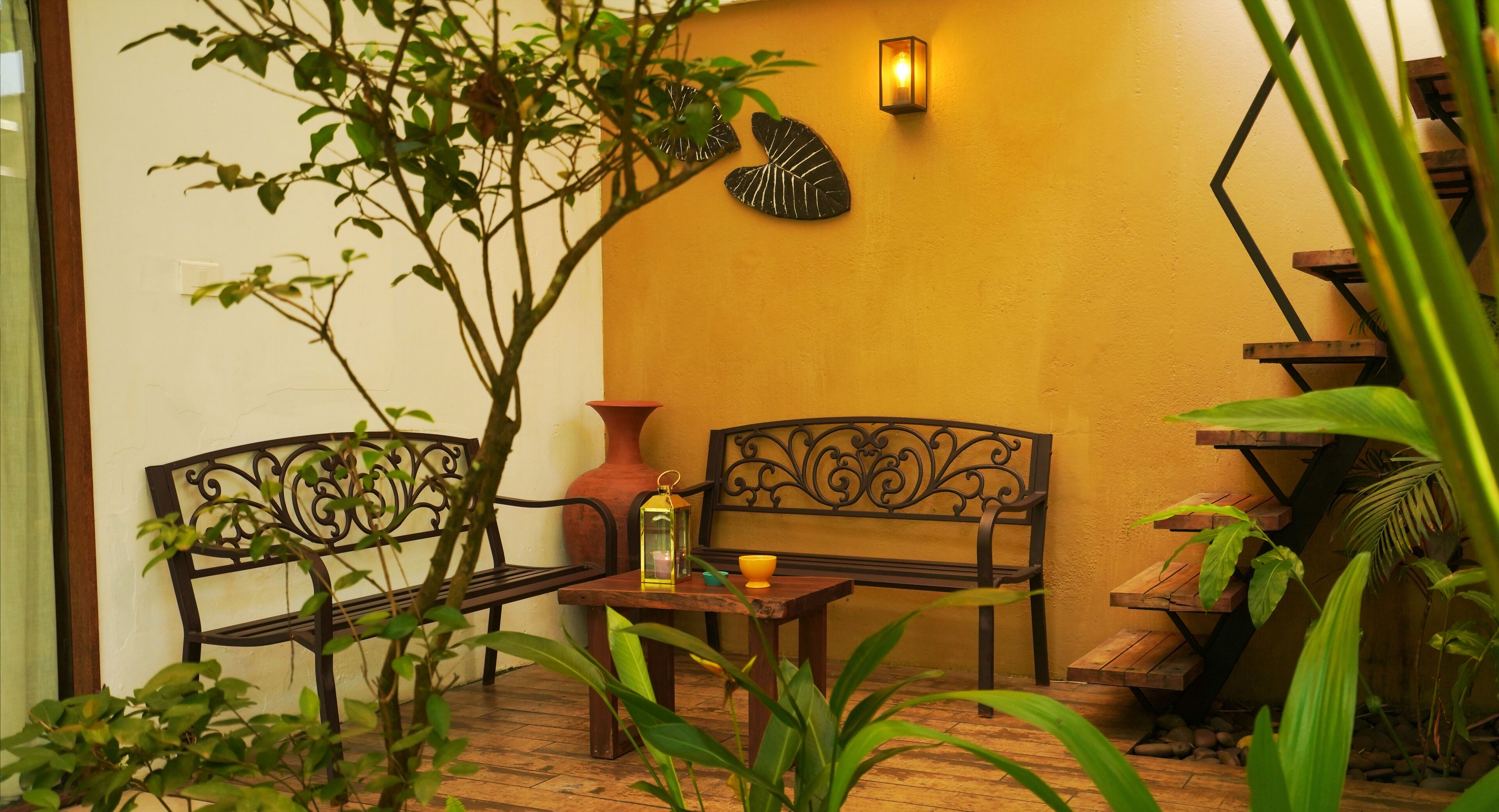 A patio with two metal benches, a small table with a lantern, and a wall-mounted light fixture.