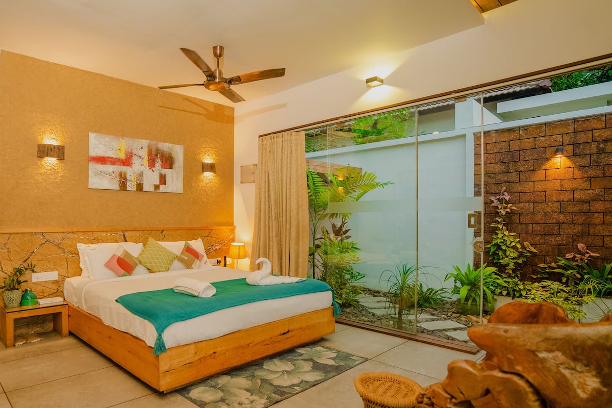 A bedroom with a bed, a TV, a ceiling fan, and a sliding glass door leading to a balcony with plants.