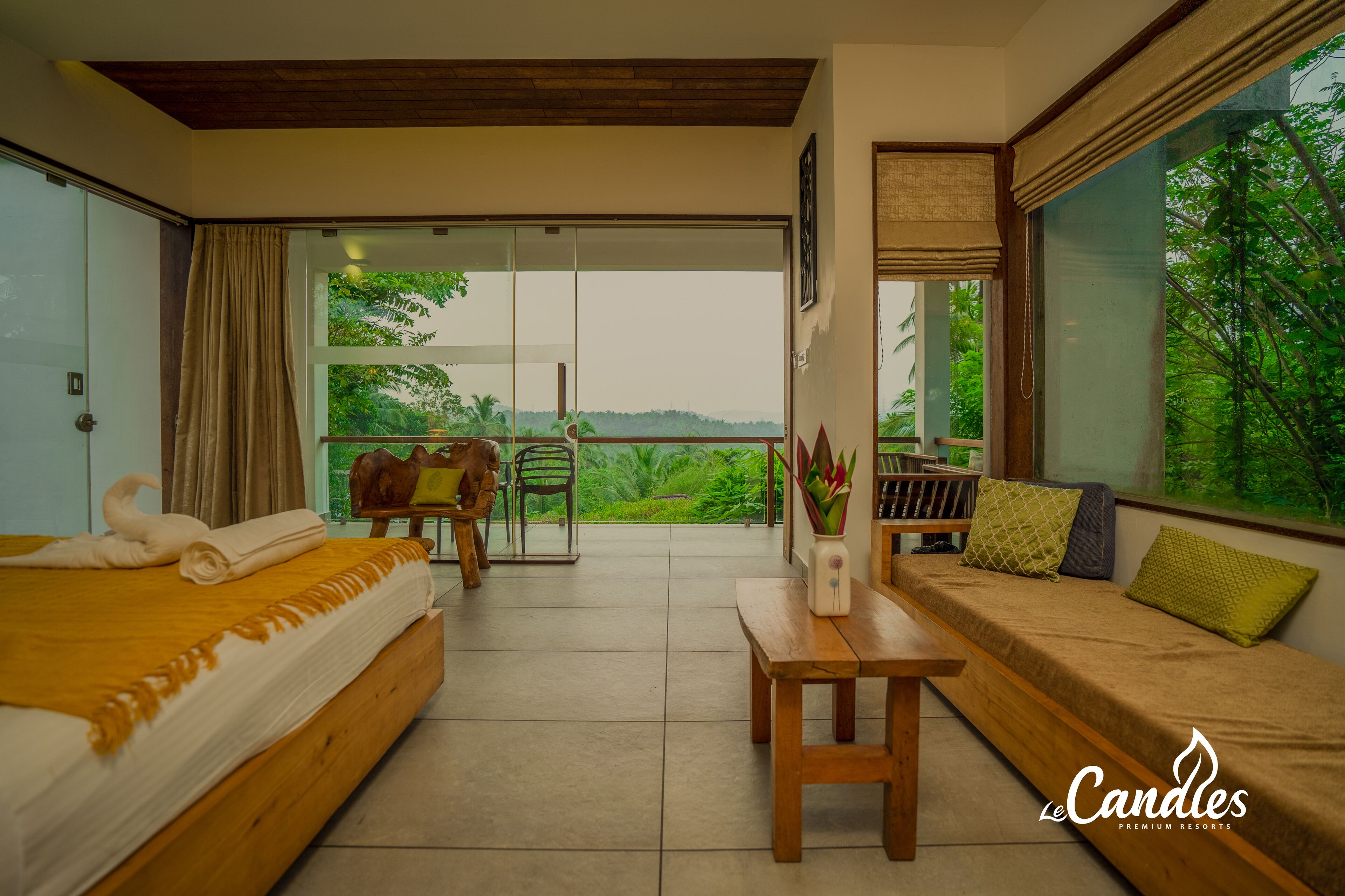 A bedroom with a bed, a sofa, a table, and a view of greenery through large windows.