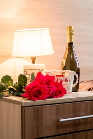 A bedside table with a bottle of champagne, a box of Confeiteria Caffello cookies, and a red rose. - Hotel Kinolenta (Vladimir)