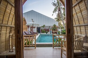 A pool area with wooden chairs and a small pool, surrounded by plants and a building with a corrugated metal exterior. - Puri Lumbung Village (Carangsari)