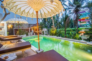 A pool area with lounge chairs and a decorative umbrella, surrounded by palm trees and a signpost with various destinations. - Puri Lumbung Village (Carangsari)