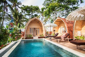 Un espace piscine agrémenté de chaises longues, de structures en bois en forme d’arc et d’une végétation tropicale.