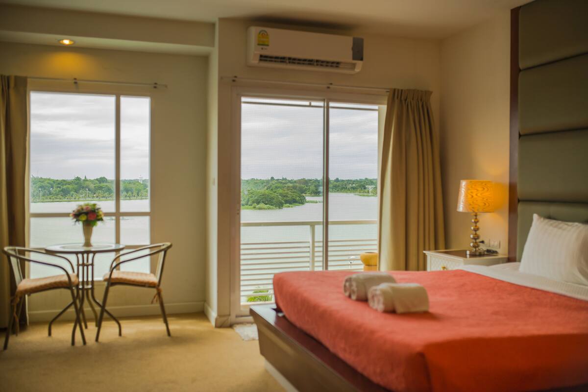 A hotel room with a bed, a table with two chairs, a window with a view of a body of water and greenery, and an air conditioning unit.