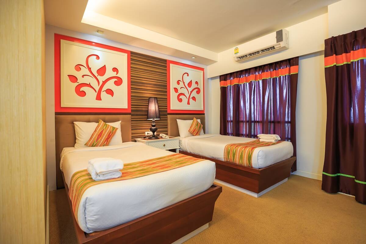 A hotel room with two beds, a nightstand, an air conditioning unit, and a window with curtains.