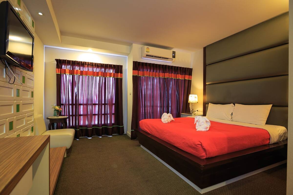 A hotel room with a bed, a desk, a chair, a TV, and an air conditioning unit.