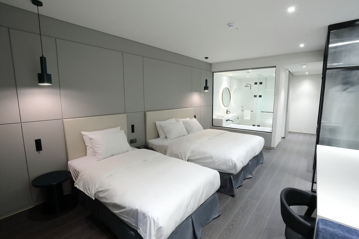 A modern hotel room with two beds, a small round table, a desk, and a bathroom with a mirror and sink.