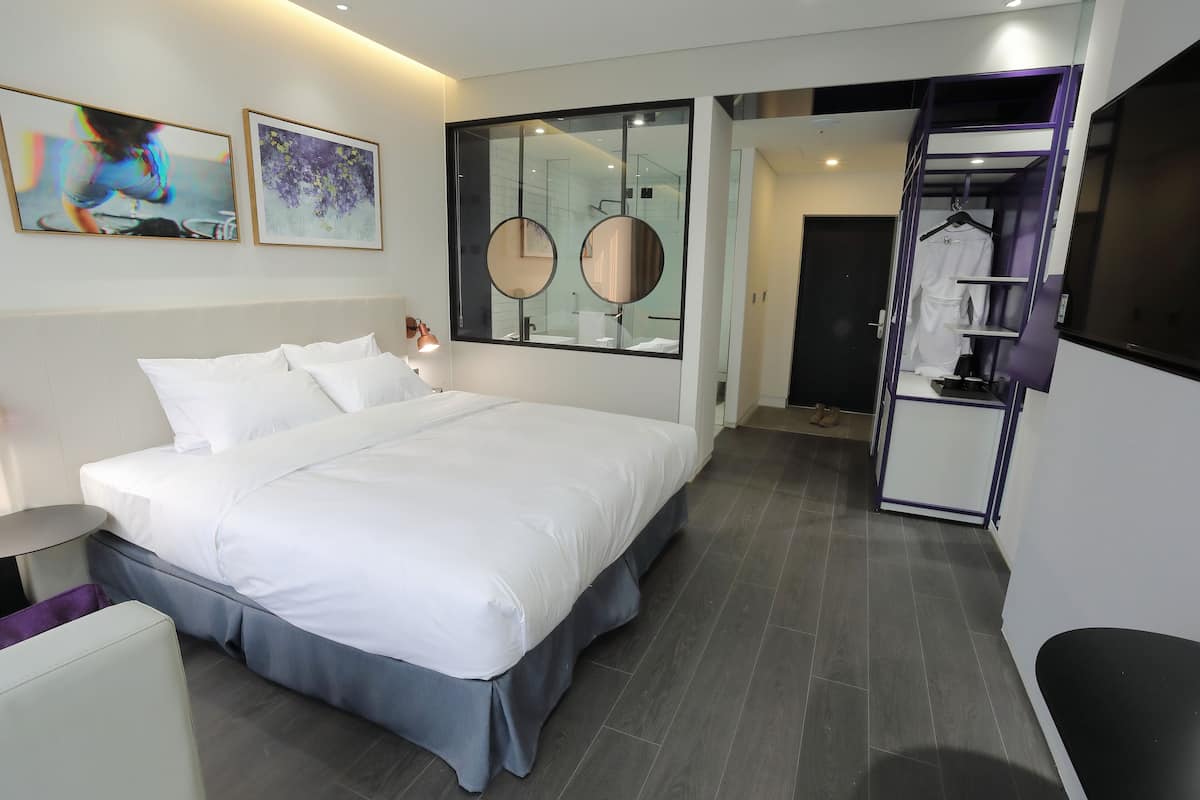 A modern hotel room with a large bed, a bedside table, a mirror, and a walk-in closet.
