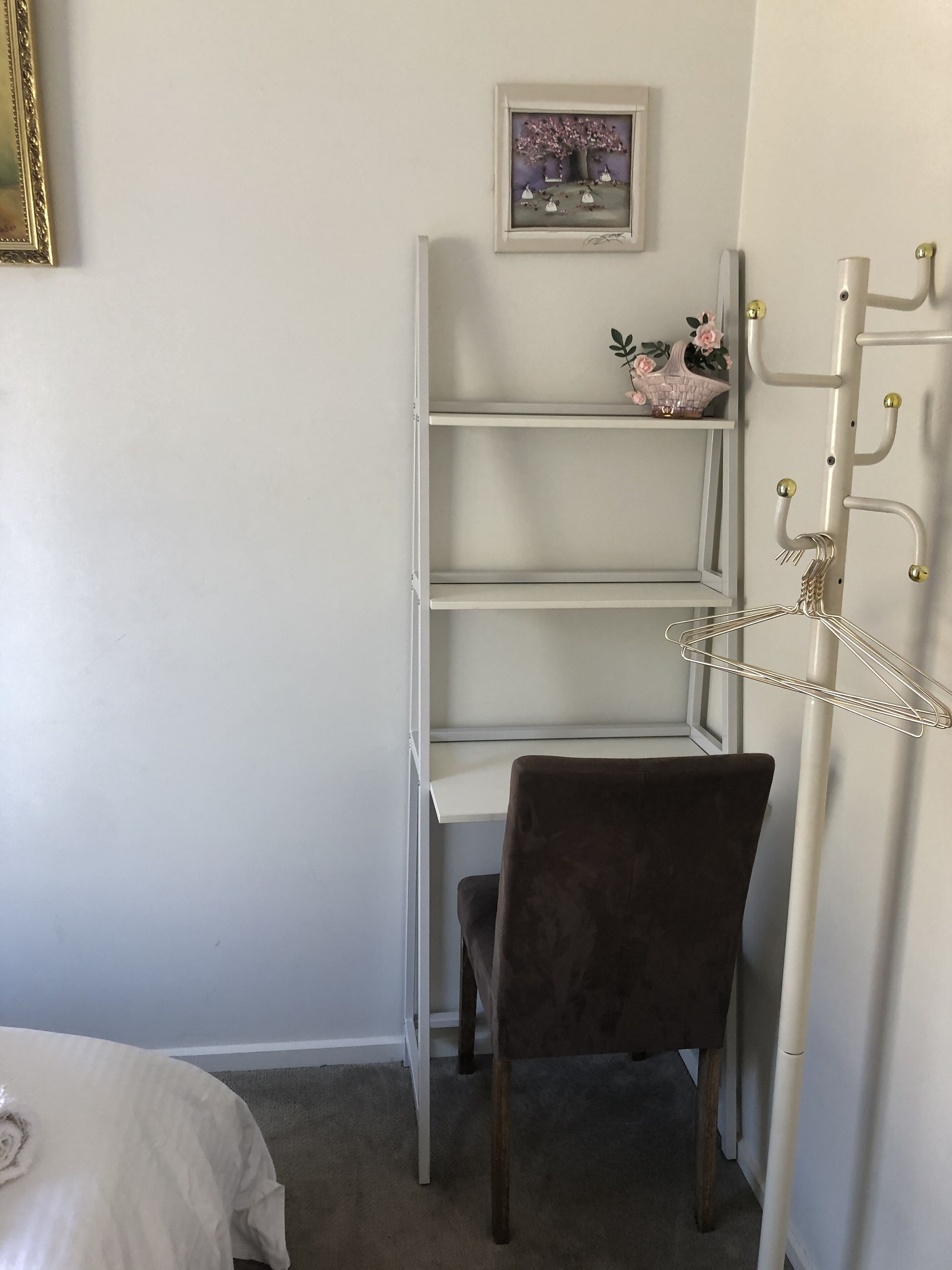 Una habitación de esquina con un estante gris, una silla marrón, un cuadro pequeño en la pared y un perchero con ganchos.