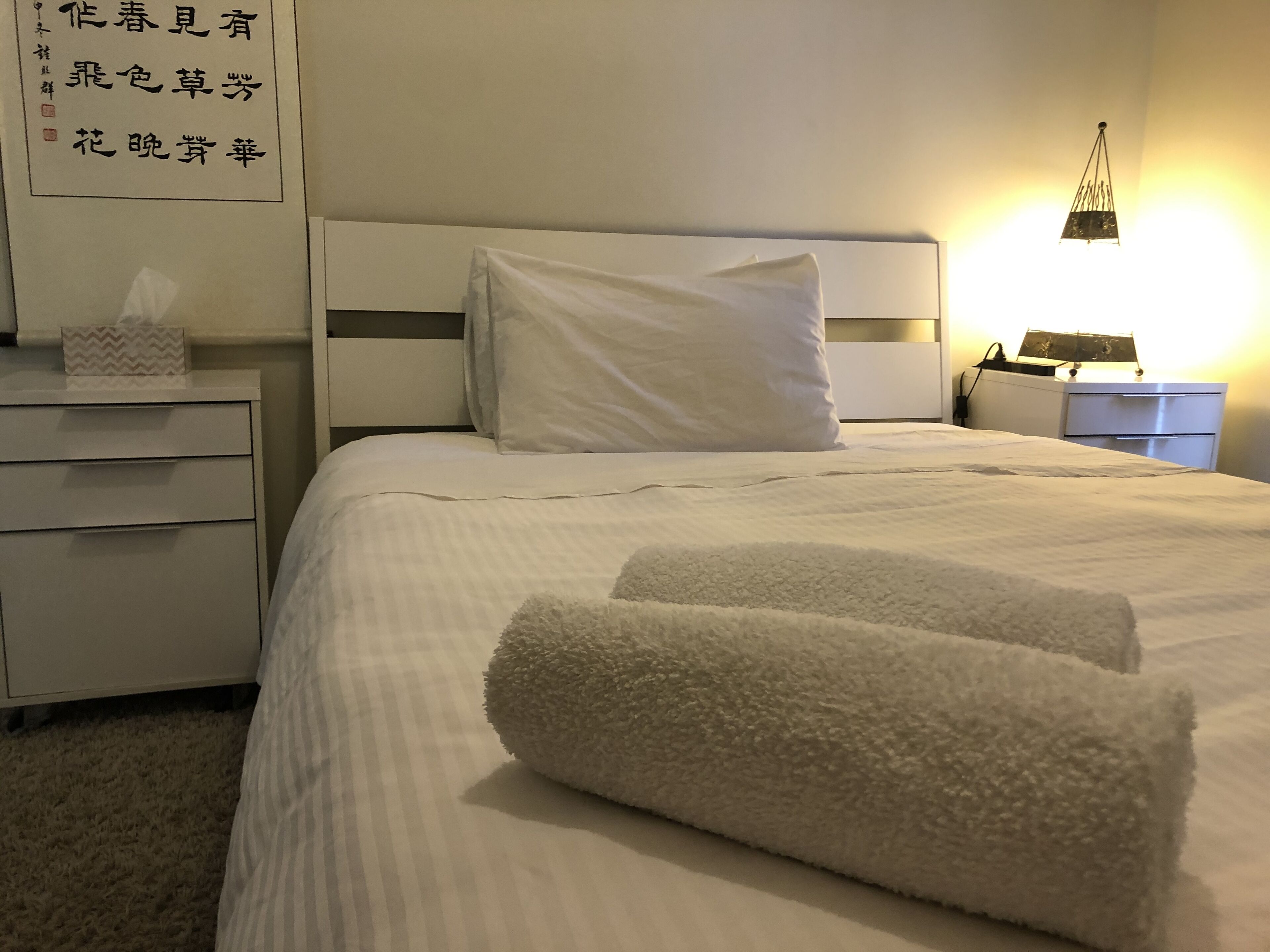 A neatly made bed with white linens and two fluffy white towels. There is a bedside table with a tissue box and a lamp. A wall-mounted shelf with decorative items is visible.