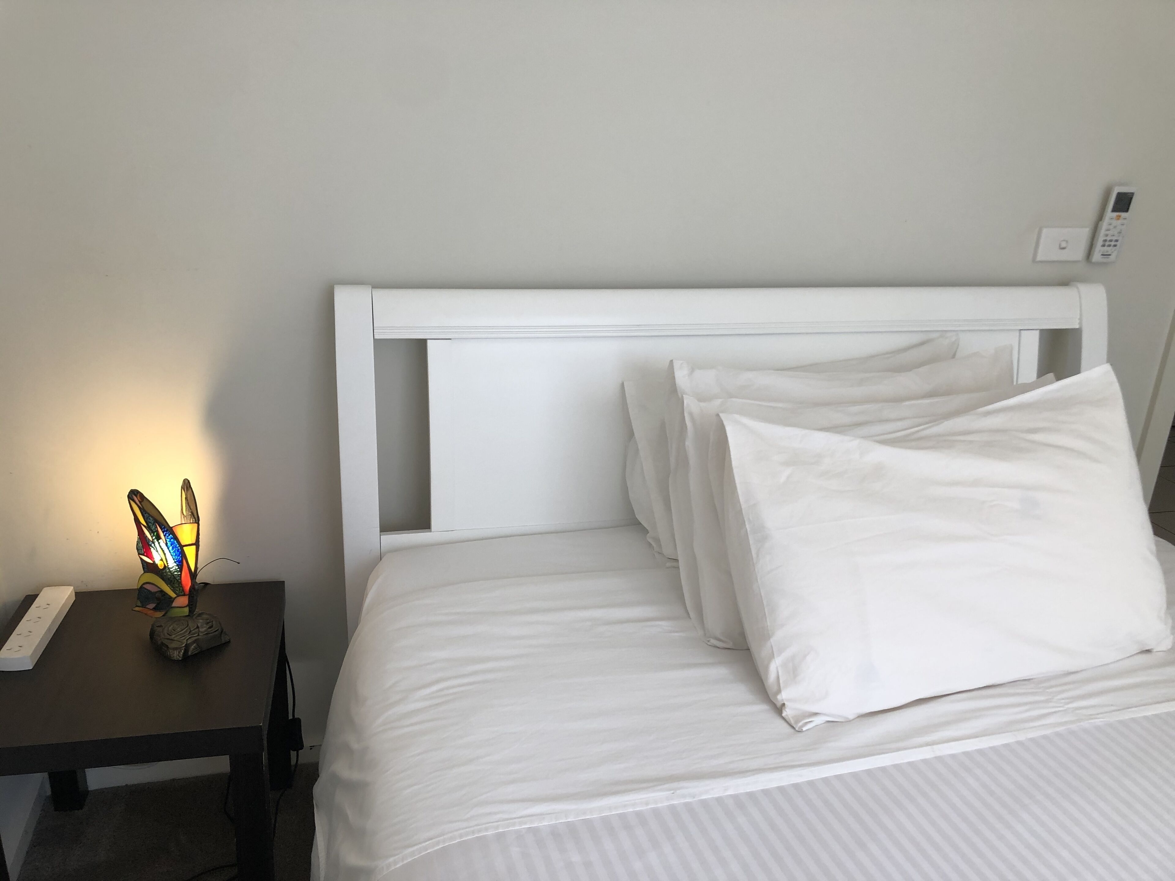 A neatly made bed with white linens and pillows, a bedside table with a decorative item, and a wall-mounted thermostat.