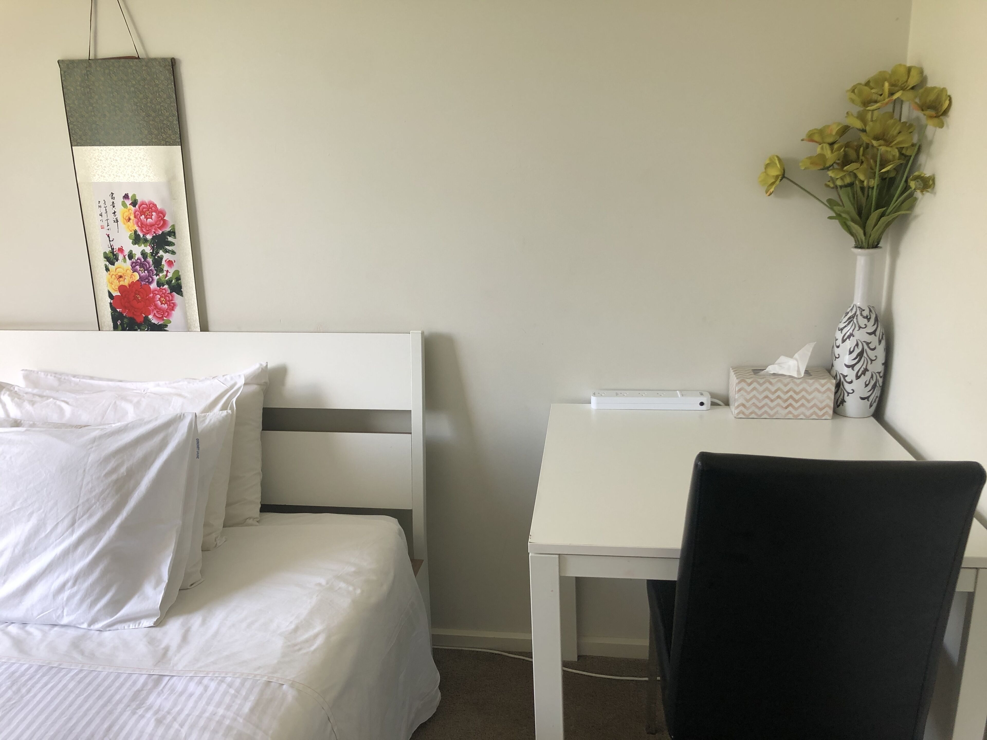 A hotel room with a bed, a desk, a chair, a vase with flowers, and a decorative item on the wall.