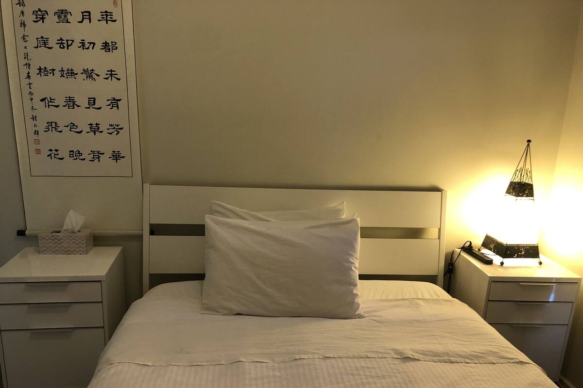 Une chambre d’hôtel avec un lit soigneusement fait, une table de chevet sur laquelle se trouve une boîte de mouchoirs, et un panneau mural orné de caractères chinois.