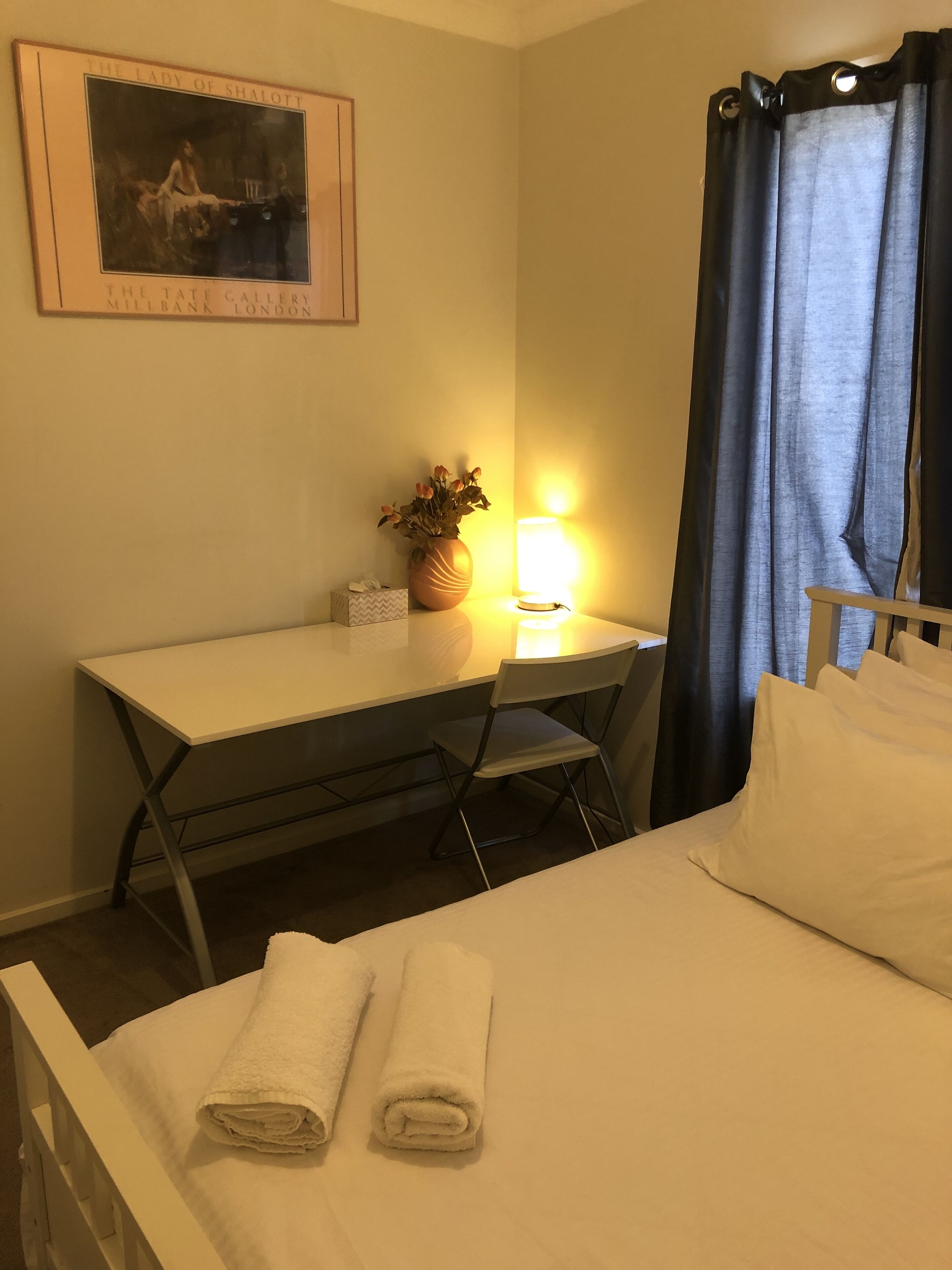 A hotel room with a bed, a desk, a chair, a lamp, a vase with flowers, and a painting on the wall.