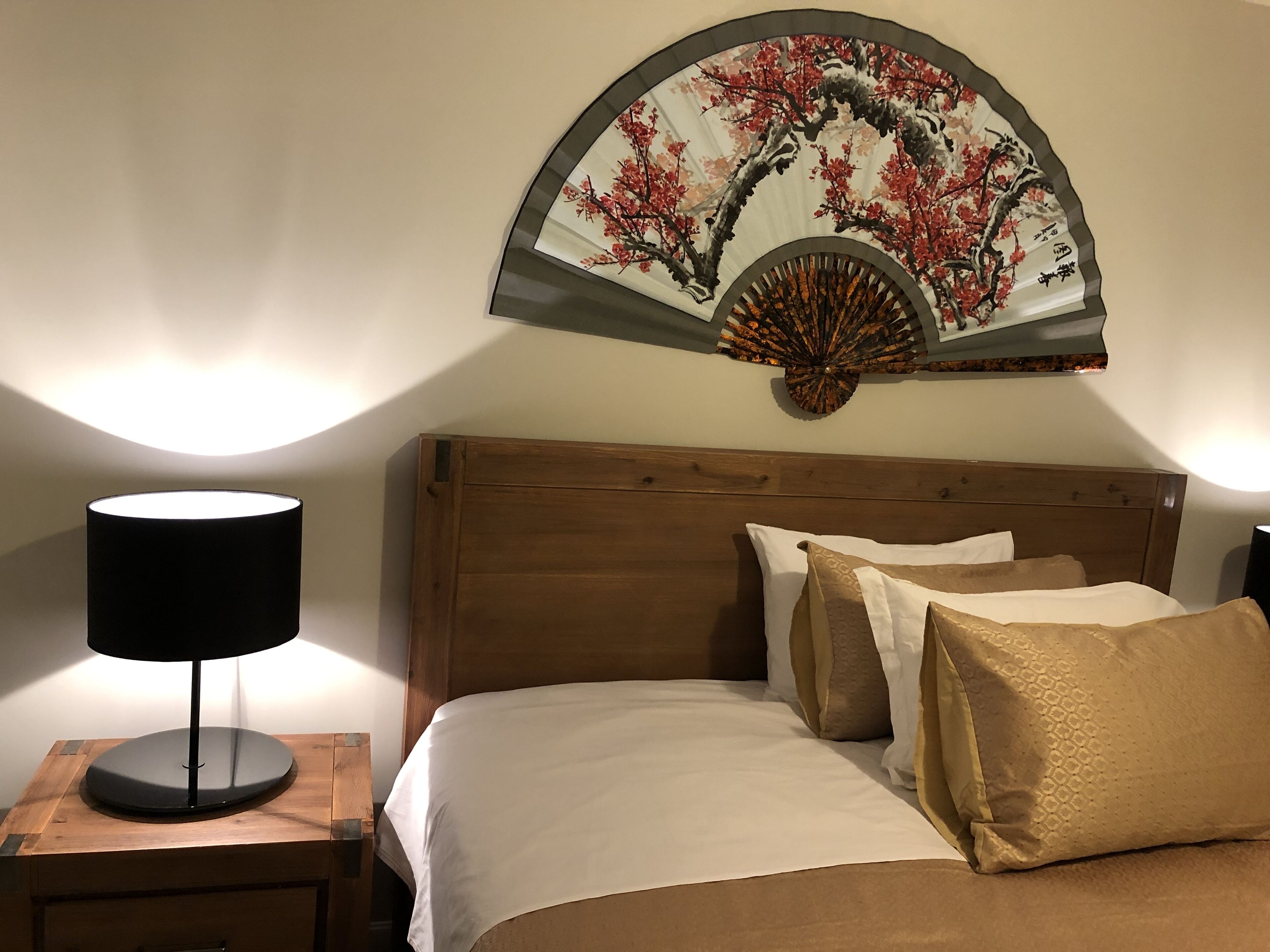 Una cama bien tendida con ropa blanca y beige, cabecera de madera y un ventilador de pared con diseño tradicional.