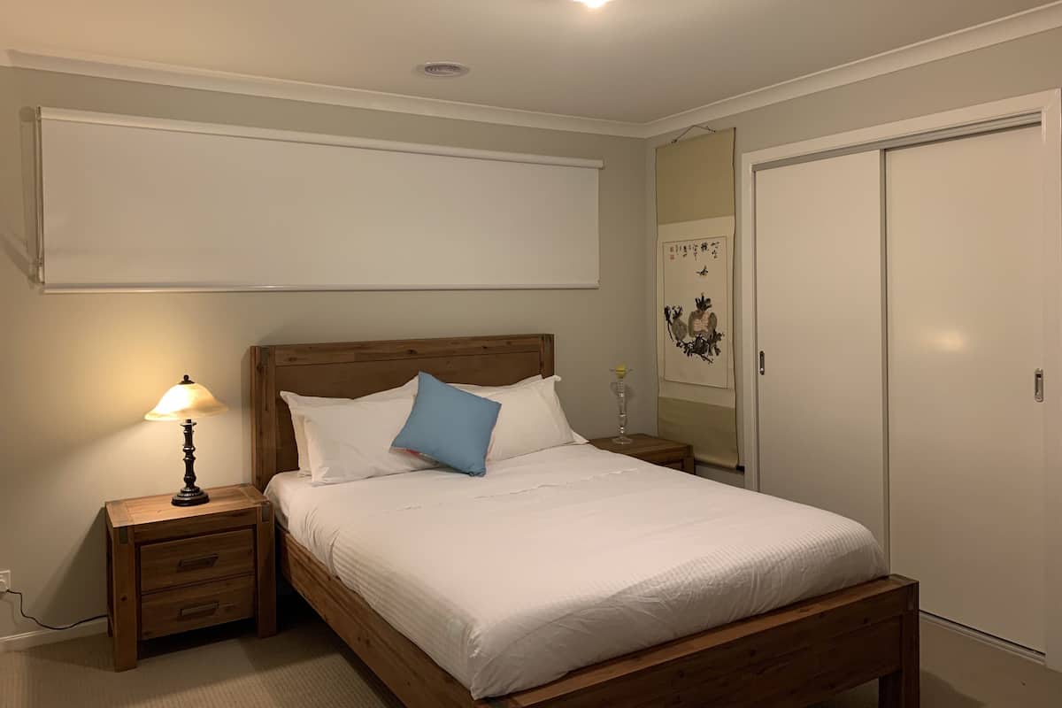 Une chambre avec un lit en bois, une literie blanche, une lampe de chevet, une table de chevet, une étagère fixée au mur et une porte dotée d’un miroir.
