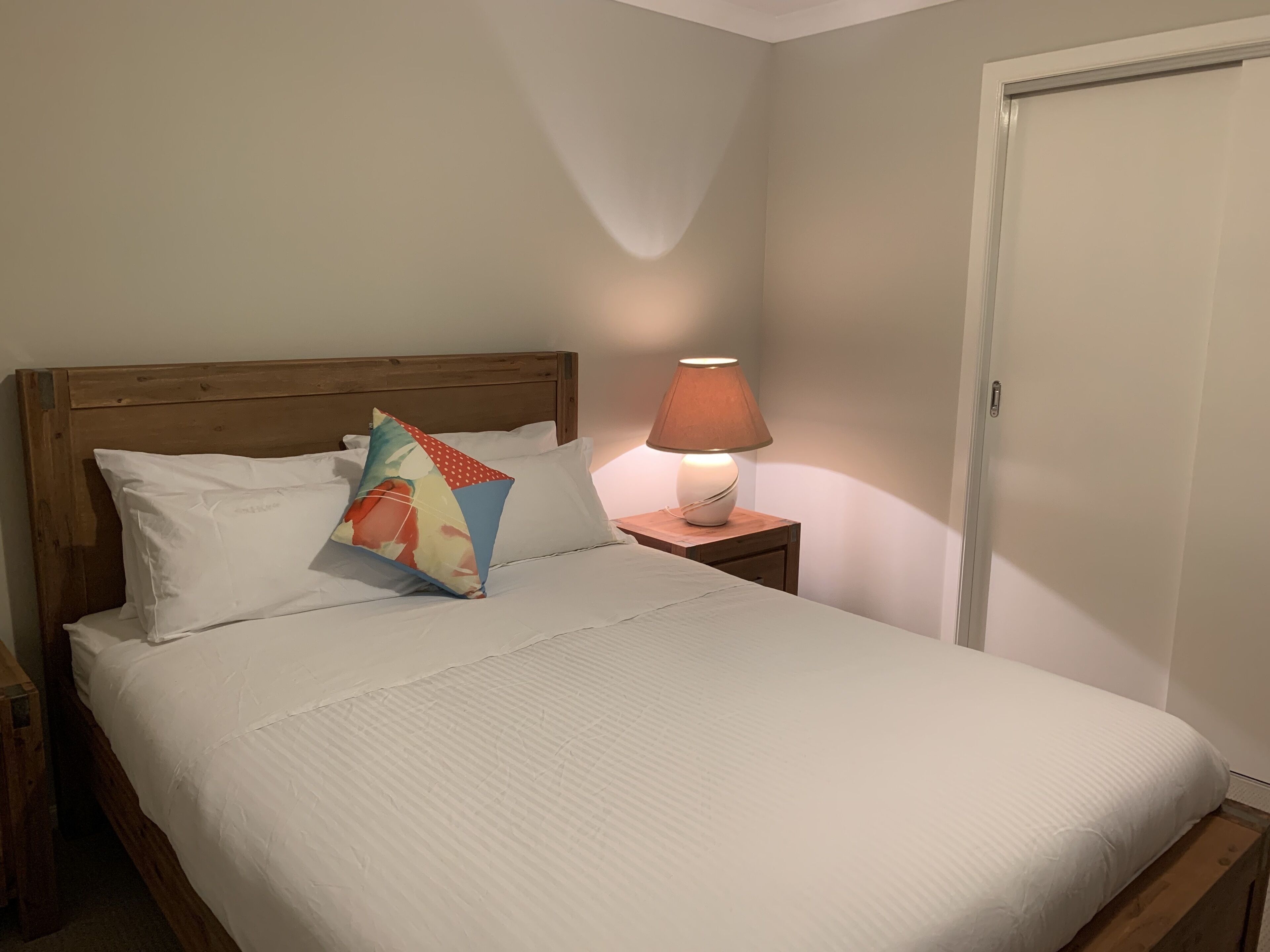 A neatly made bed with white bedding and a colorful pillow, a wooden headboard, a bedside lamp, and a closed door in a simple room.