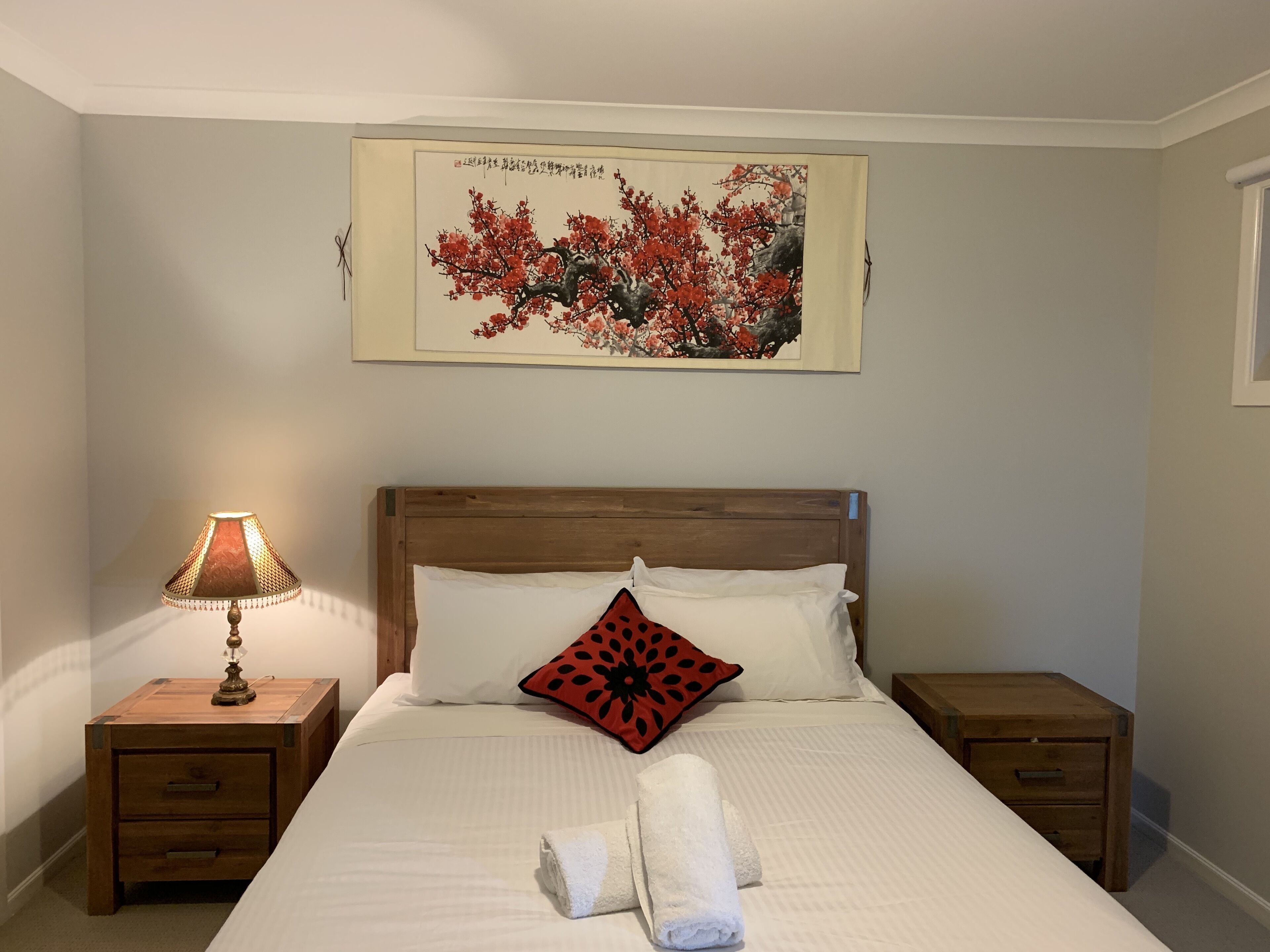 Een net opgemaakt bed met witte lakens, een kussen met rode bloemen en een witte handdoek. Links staat een houten nachtkastje met een lamp, en rechts een ander nachtkastje. Daarboven hangt een kunstwerk in een lijst.