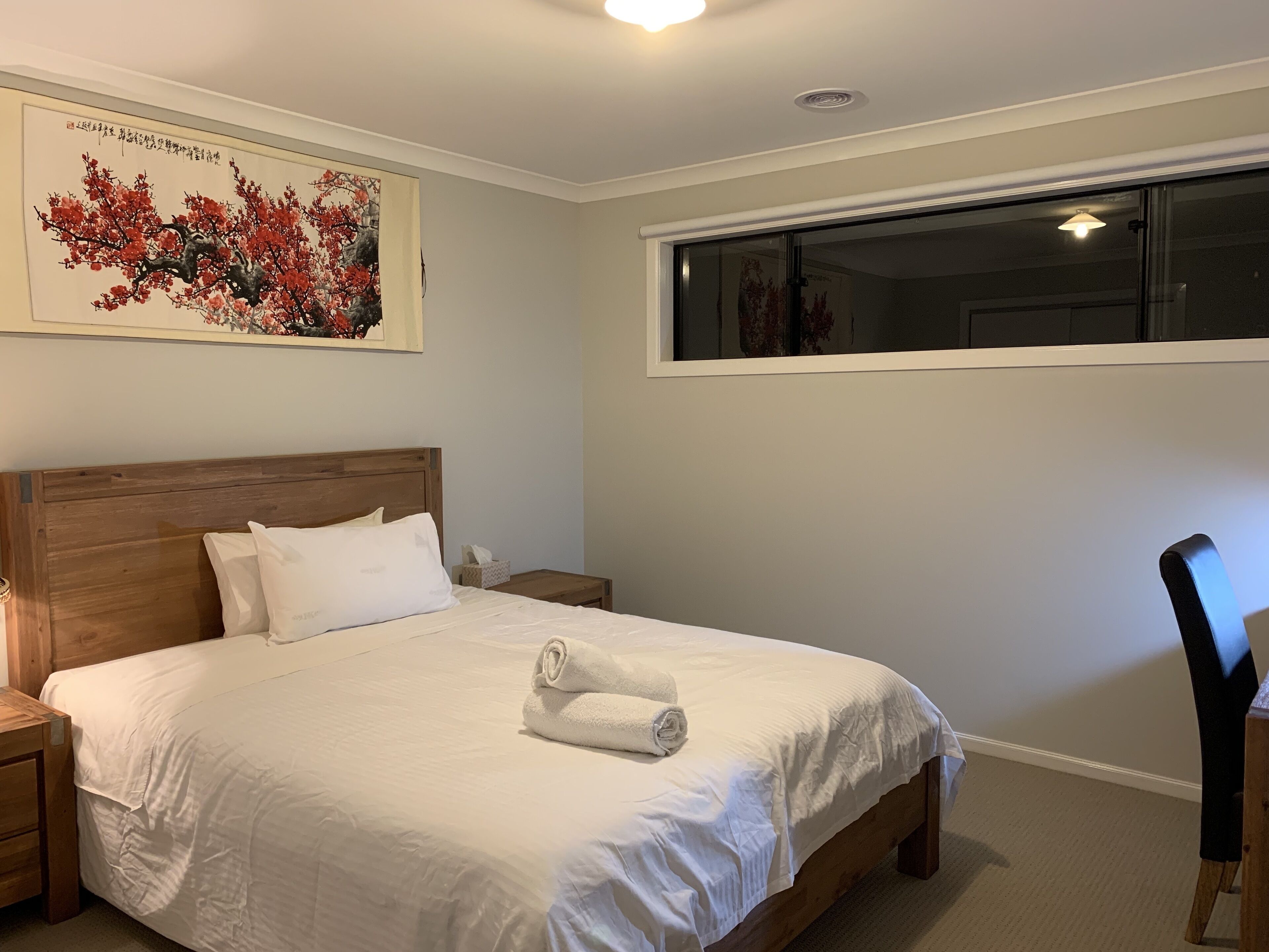 A neatly made bed with white linens, a wooden headboard, and a rolled towel on the bed. There is a nightstand with a lamp and a tissue box beside the bed. A large painting of red flowers hangs on the wall above the bed.